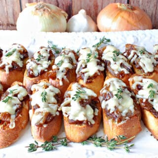 A horizontal photo of a white platter filled with French Onion Crostini with melted cheese and thyme sprigs on top.