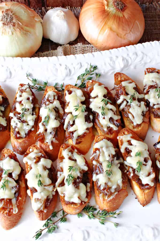 An overhead vertical photo of a tray filled with French Onion Crostini along with onions and garlic in the background.