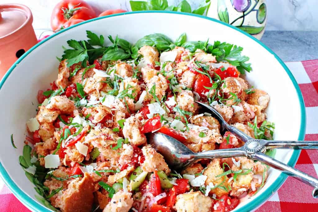 A horizontal closeup of an Italian Panzanella Salad in a large serving bowl with tongs, fresh basil, tomatoes, and Parmesan cheese.