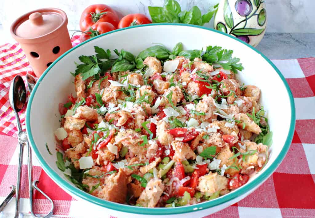 A horizontal closeup of a large bowl of Italian Panzanella Salad on a red and white tablecloth with salad tongs on the side.