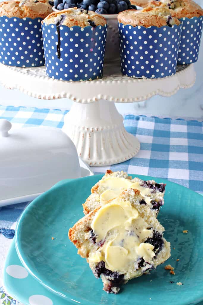 A vertical closeup of a NY Times Blueberry Muffin on a blue plate with butter along with a cake stand filled with muffins in the background.