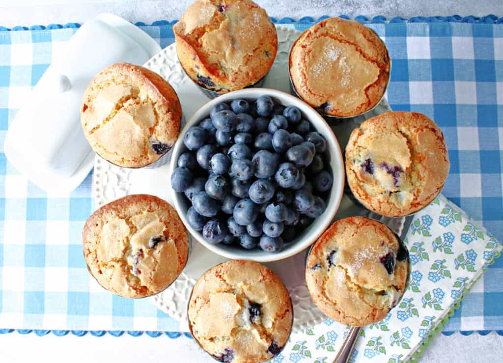 A direct overhead photo of a round cake plate filled with NY Times Blueberry Muffins along with a bowl of fresh blueberries in the center.