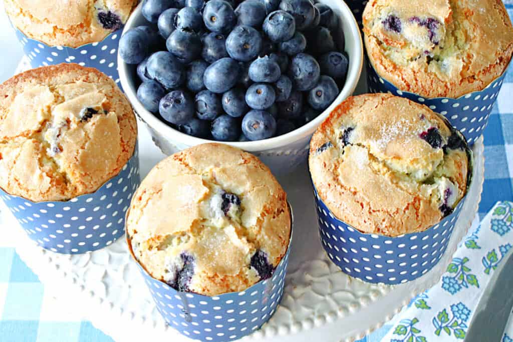 A horizontal overhead closeup photo of some NY Times Blueberry Muffins on a plate with fresh blueberries in the center and a blue and white checked napkin.