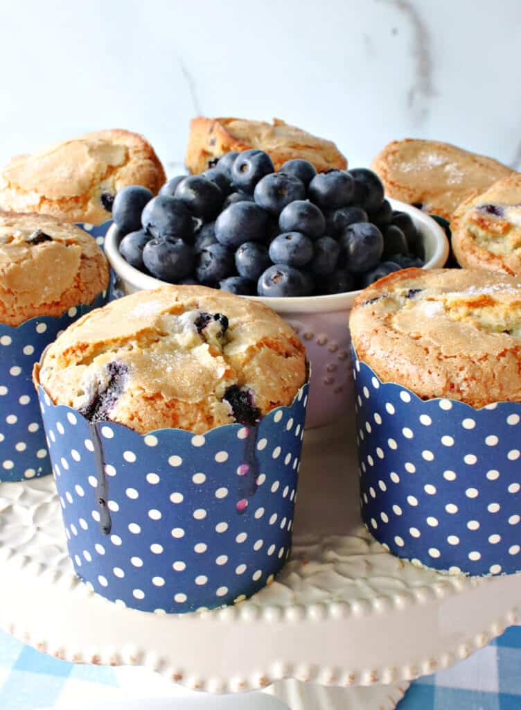 A closeup vertical photo of some NY Times Blueberry muffins on a cake stand with a bowl of fresh blueberries in the center.