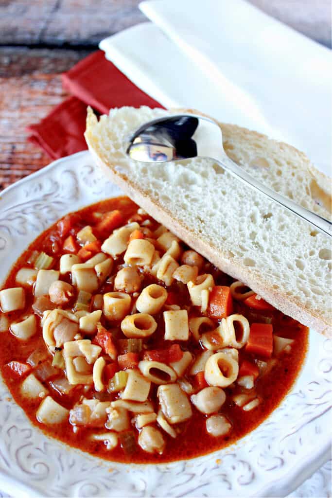 An overhead vertical closeup of a white bowl filled with Tuscan Pasta Soup with a spoon and a slice of bread on the side.
