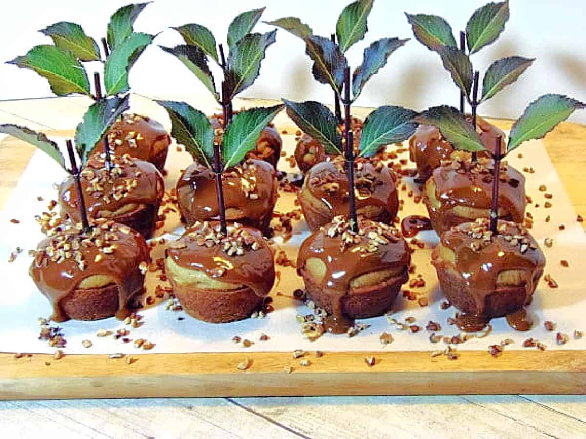 Twelve caramel coated Caramel Apple Cupcakes on a wooden board with leaves and nuts.