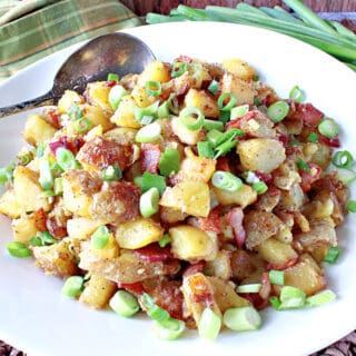 A white bowl filled with German Fried Potatoes along with scallions and a spoon.