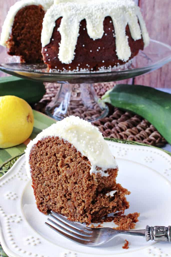 A vertical closeup of a slice of Gingerbread Bundt Cake with Zucchini on a plate with an entire cake on a cake stand in the background.