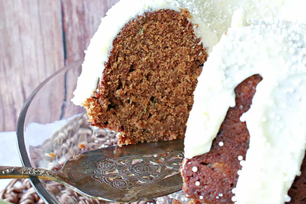 A Gingerbread Bundt Cake with Zucchini on a glass cake stand with cake server and a slice taken out.