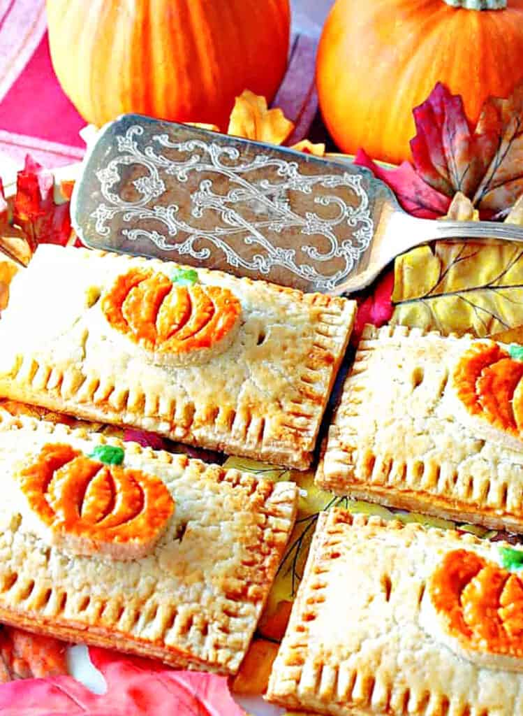 A vertical overhead closeup photo of 4 Pumpkin Hand Pies on a platter with a silver serving spatula and autumn leaves.