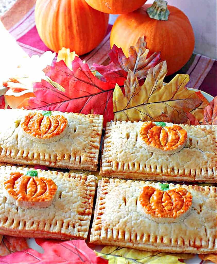 A vertical closeup image of four Pumpkin Hand Pies with pumpkin pie crust embellishments on a platter with autumn leaves and pumpkins in the background.