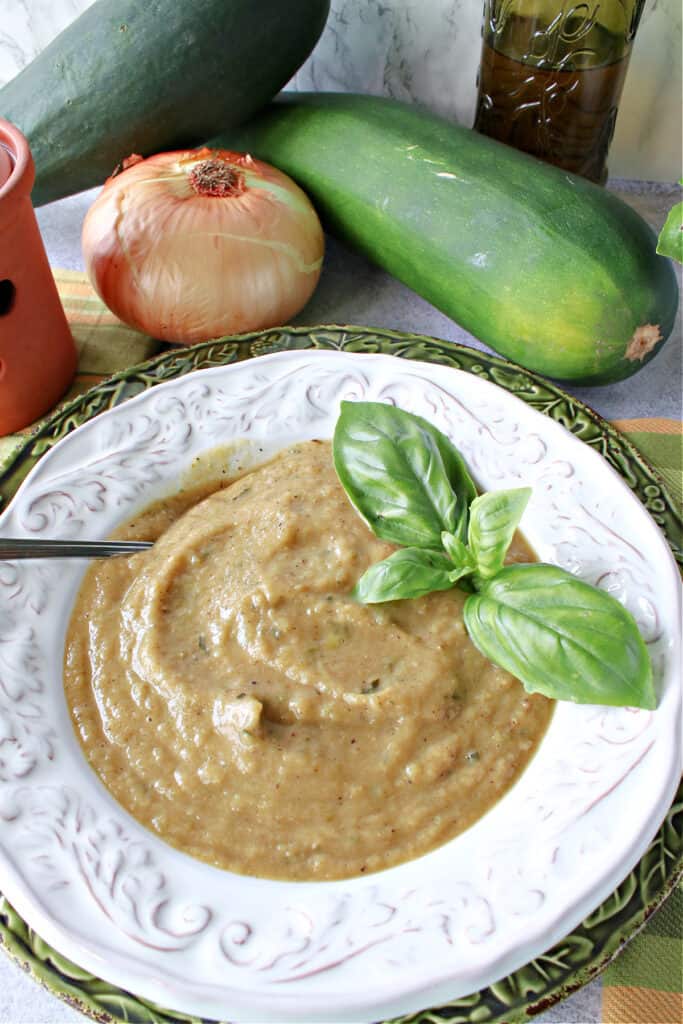 A vertical overhead closeup of a creamy bowl of Roasted Garlic and Zucchini Soup with fresh basil as garnish with a spoon in the bowl.