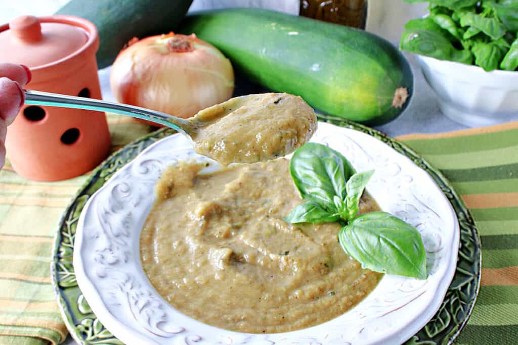 A spoonful of Roasted Garlic and Zucchini Soup over a white bowl with fresh basil as garnish and an onion and zucchini in the background.