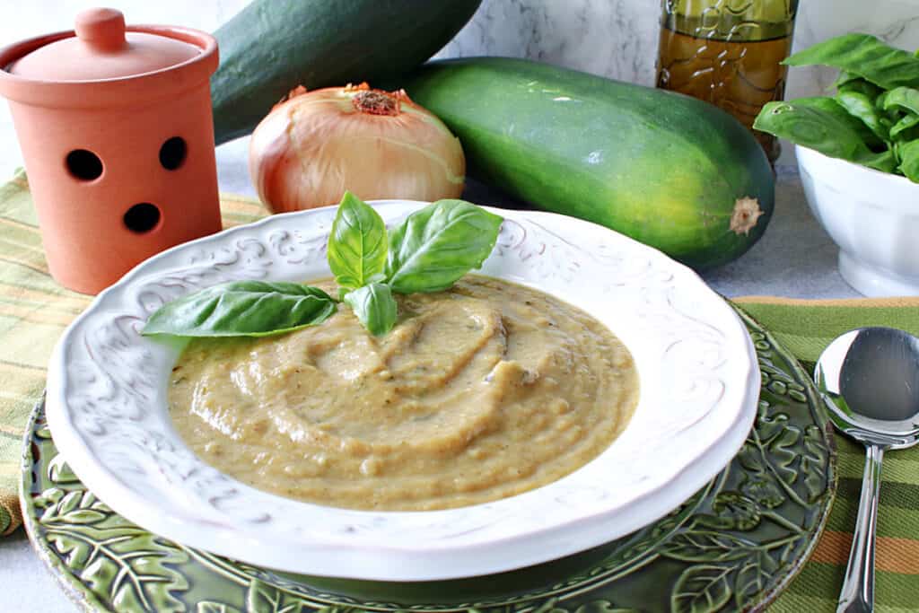 A white bowl filled with Roasted Garlic and Zucchini Soup along with some fresh basil and a spoon on the side.