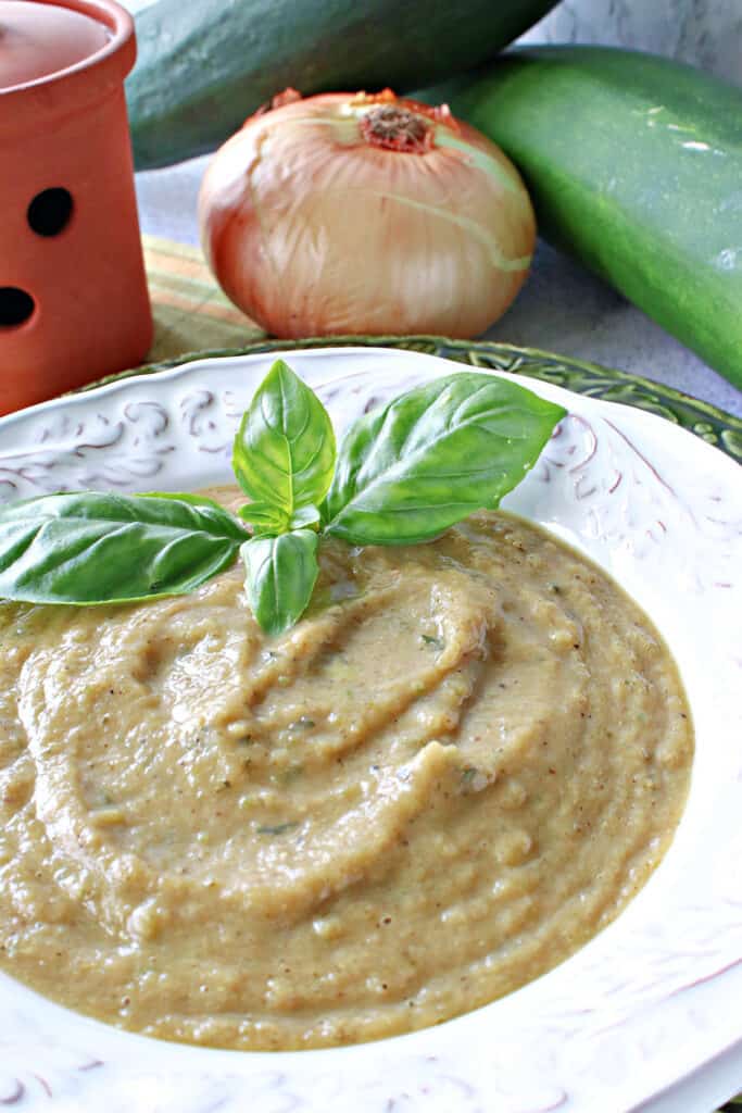 A vertical closeup of a bowl of Roasted Garlic and Zucchini Soup along with fresh basil leaves and an onion and fresh zucchini in the background.