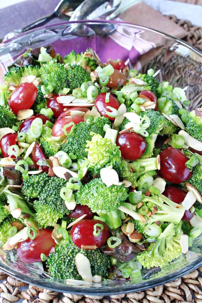 A closeup vertical image of a Broccoli Salad with Sweet and Sour Dressing in a glass bowl with red grapes and slivered almonds.