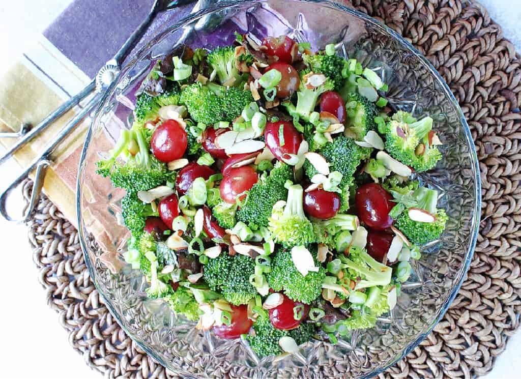 A direct overhead photo of a Broccoli Salad in a glass bowl with salad tongs and a colorful napkin on the side.