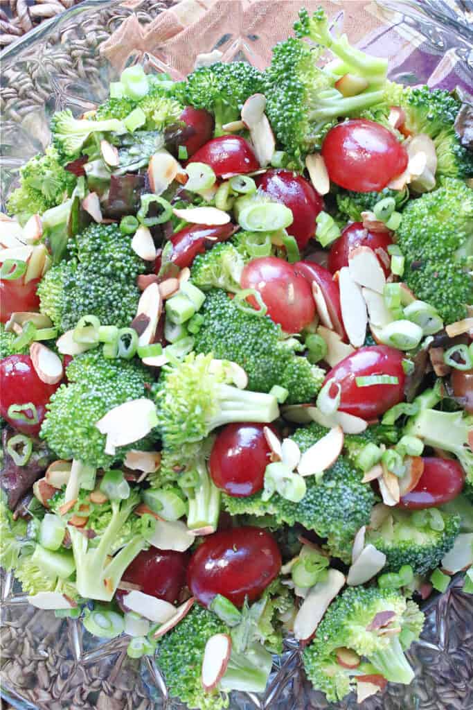 A vertical closeup of a fresh Broccoli Salad in a glass bowl with scallions, almonds, red leaf lettuce, and grapes.