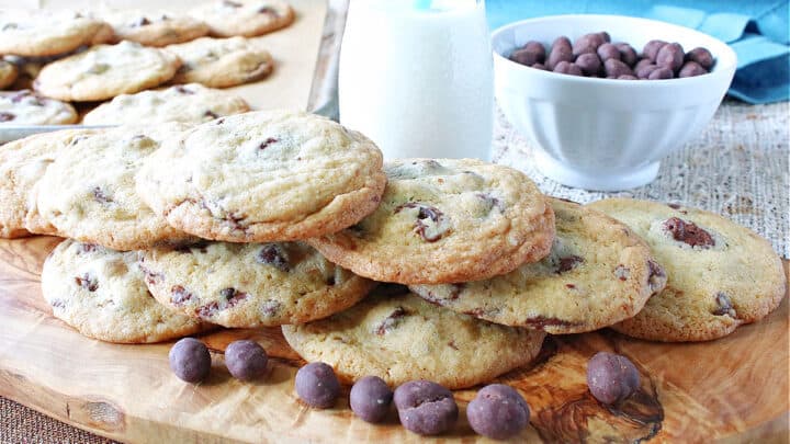 A photo of a stack of Chocolate Chip Espresso Cookies on a wooden board with a glass of milk and a blow of chocolate chips in the background.