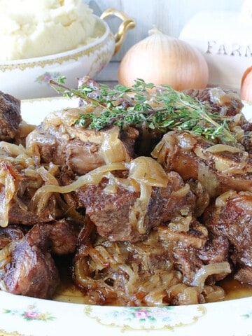 A platter filled with French Onion Short Ribs with fresh thyme sprigs on top and a bowl of mashed potatoes in the background.