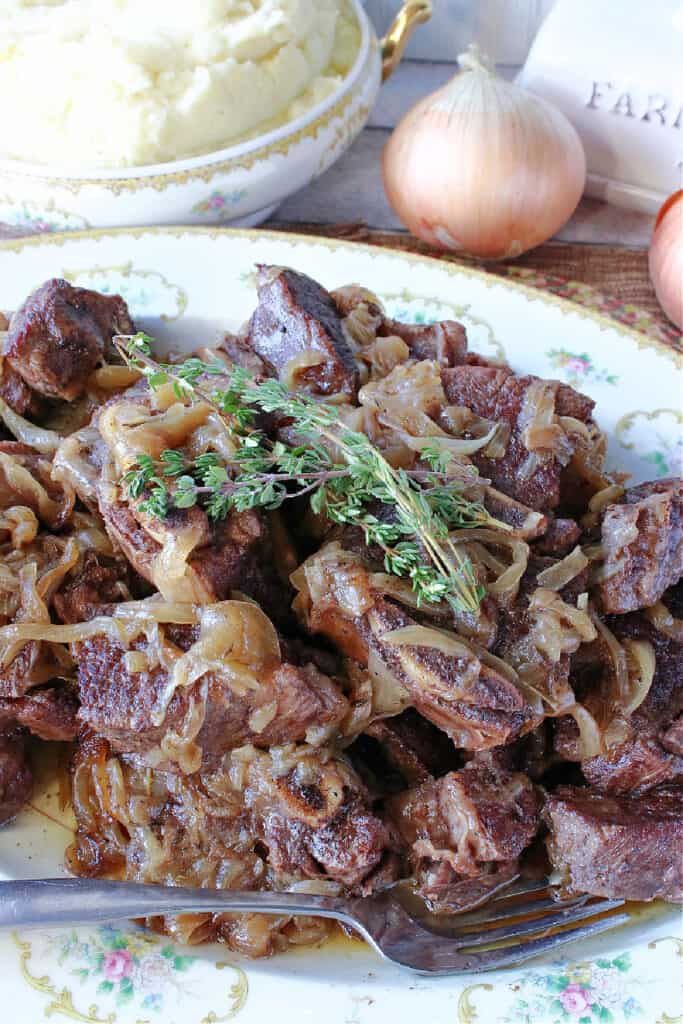 A vertical closeup of a platter of French Onion Short Ribs with a bowl of mashed potatoes in the background.