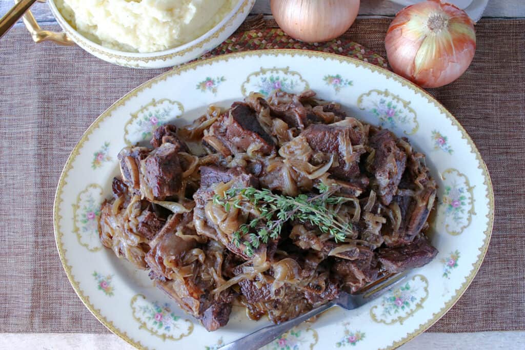 A horizontal overhead photo of a platter filled with French Onion Short Ribs with caramelized onions, fresh thyme and a fork.