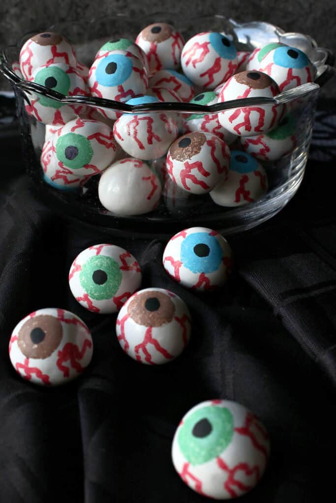A vertical closeup of a glass bowl of Gumball Eyeballs in the background and some gumball eyeballs in the front on a black napkin.