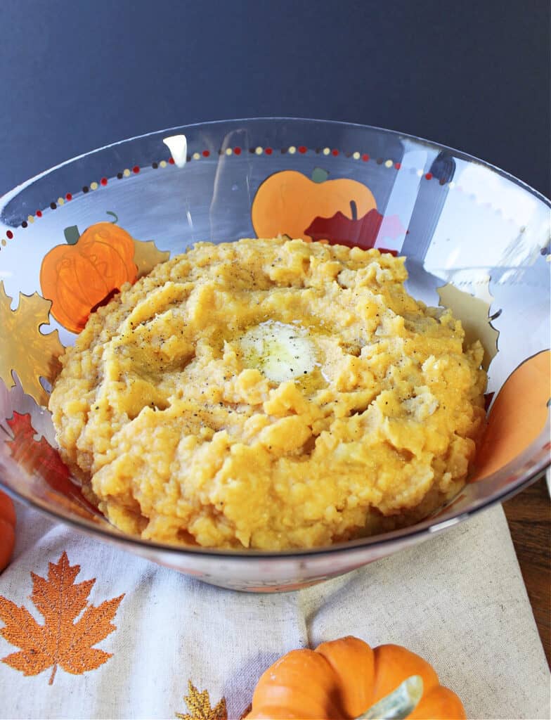 A vertical closeup of a glass bowl filled with Pumpkin Mashed Potatoes along with melted butter and black pepper on top.