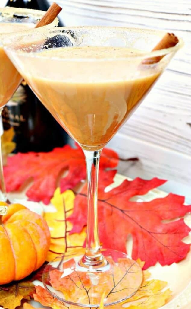A vertical closeup photo of a Pumpkin Spice Martini with a cinnamon stick and  fall leaves on the table.