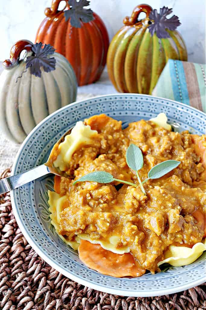 An offset vertical closeup of a blue bowl filled with Ravioli with Pumpkin Vodka Sauce along with sausage and fresh sage as a garnish.