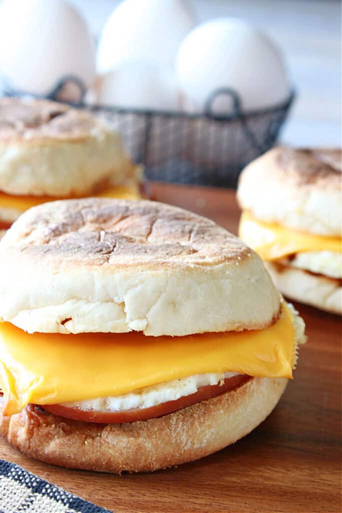A vertical closeup of a Copycat Egg McMuffin Sandwich in the foreground and a small basket of eggs in the background.