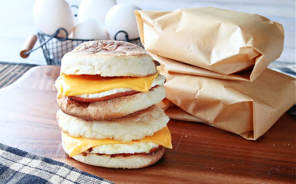 Two Copycat Egg McMuffin Sandwiches stacked on a wooden board with wrapped sandwiches in the background.