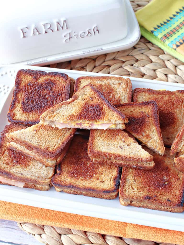 An overhead vertical picture of a tray of Grilled Ham and Cheese Appetizers on a rattan place mat along with a white butter dish.