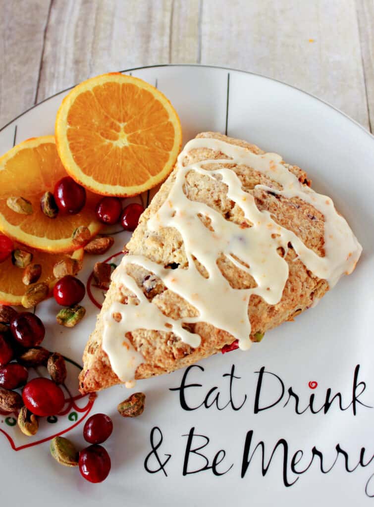 A vertical closeup of a glazed Cranberry Orange Scone on a plate with orange slices, cranberries, and pistachios as garnish.