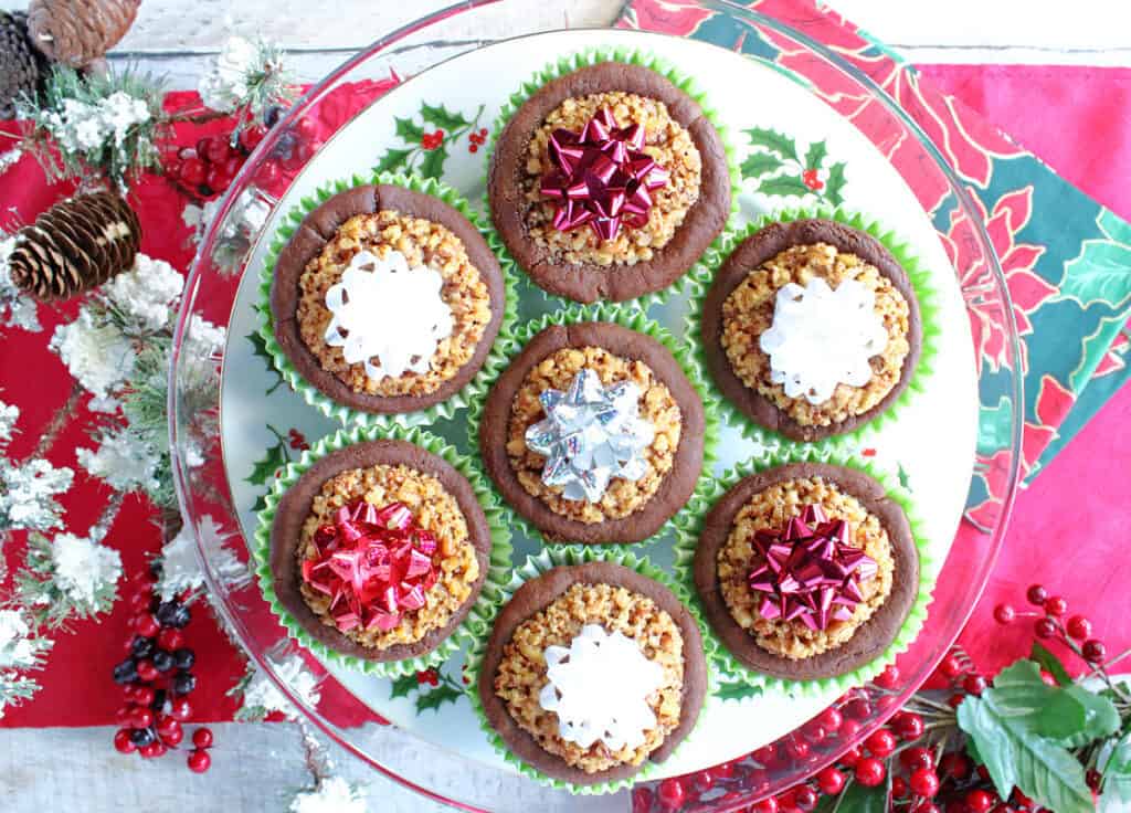 A horizontal overhead photo of a plate of Gingerbread Pecan Pie Tarts on a red table runner with holly berries and snow branches.