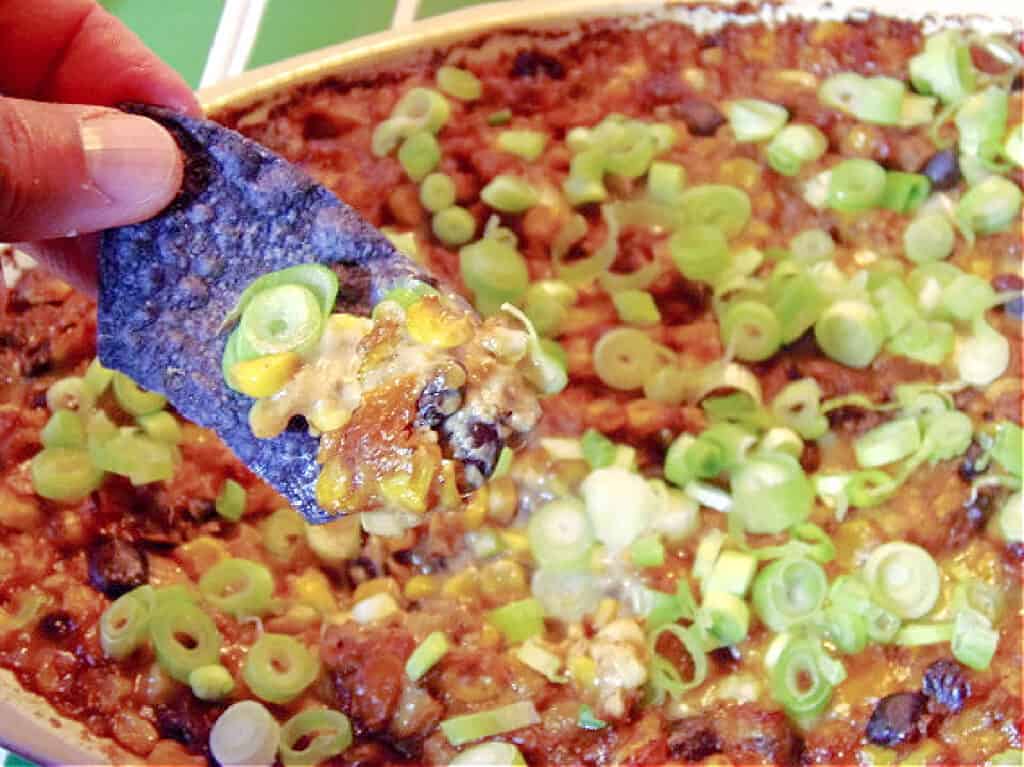 A closeup horizontal photo of a hand holding a blue tortilla chip that's been dipped into Cheesy Corn and Black Bean Dip.