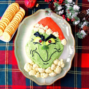 A fun Grinch Guacamole on a platter with crackers and some Christmas ornaments in the background.