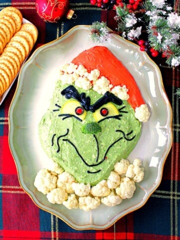 A fun Grinch Guacamole on a platter with crackers and some Christmas ornaments in the background.