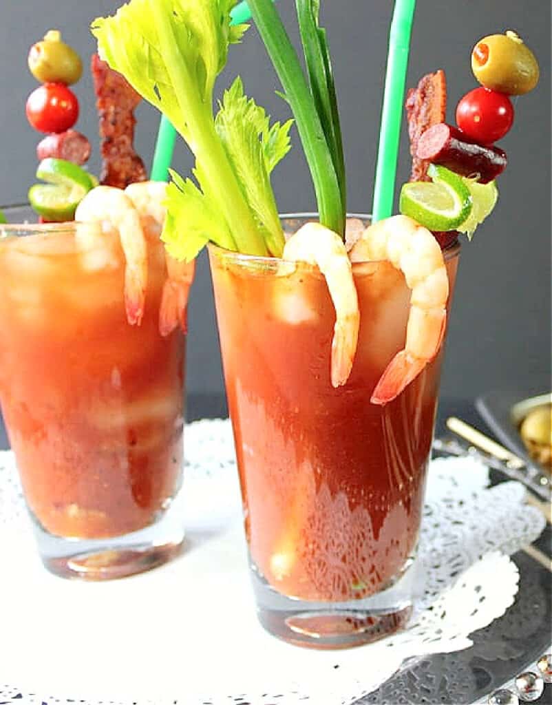 A close up vertical image of a Bacon Bloody Mary in a tall glass with celery, shrimp, lime, sausage, bacon and olives.