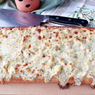 A loaf of Chicken Alfredo Garlic Bread Pizza on a wooden cutting board with a cleaver in the background.
