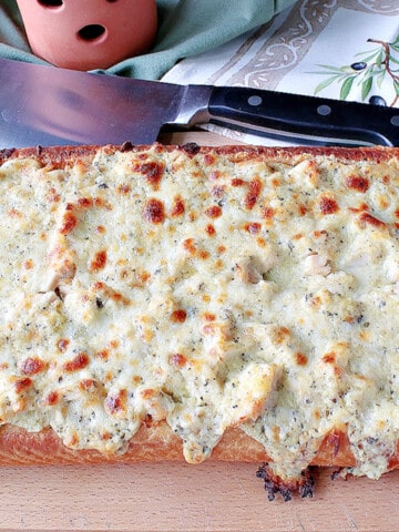 A loaf of Chicken Alfredo Garlic Bread Pizza on a wooden cutting board with a cleaver in the background.