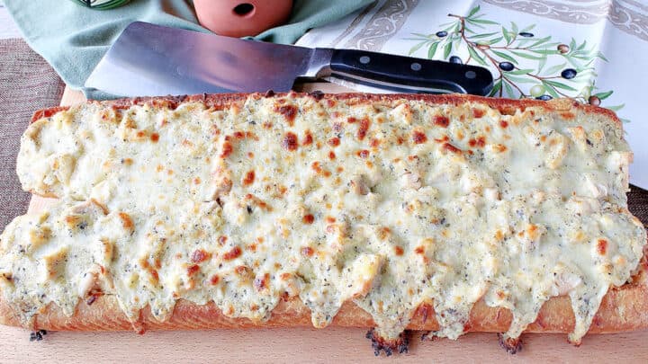 A loaf of Chicken Alfredo Garlic Bread Pizza on a wooden cutting board with a cleaver in the background.