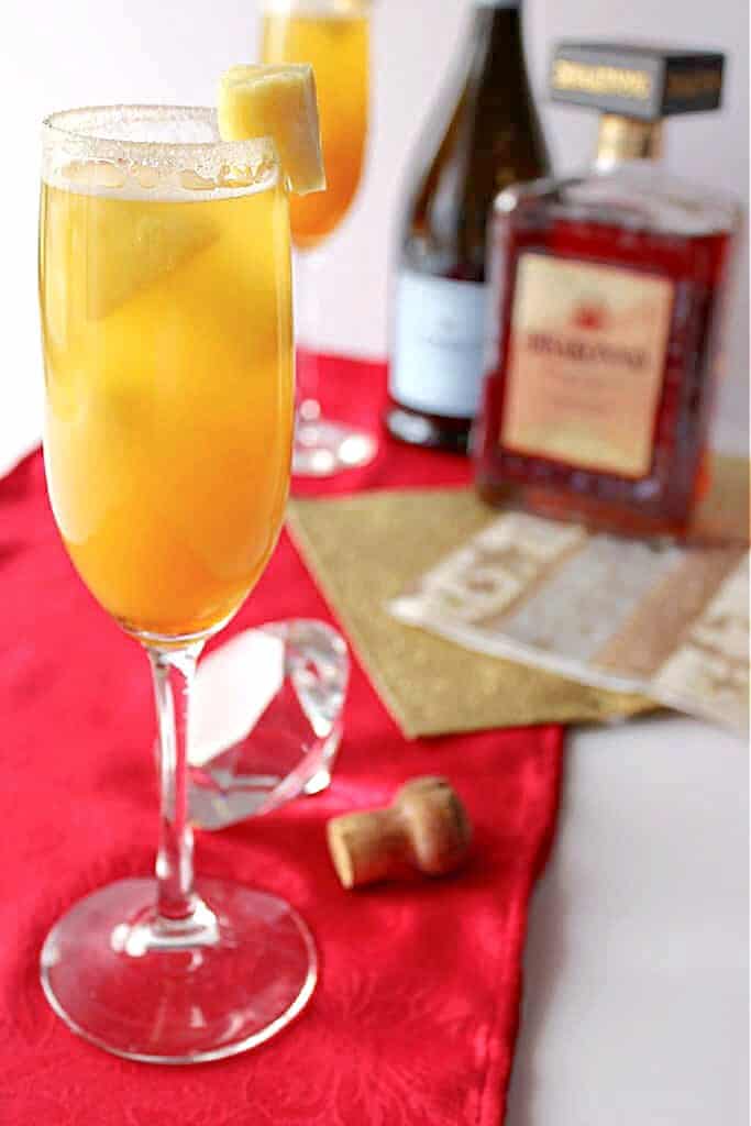A closeup vertical image of a Golden Statue Prosecco Cocktail with pineapple in the foreground and a bottle of prosecco and amaretto in the background.