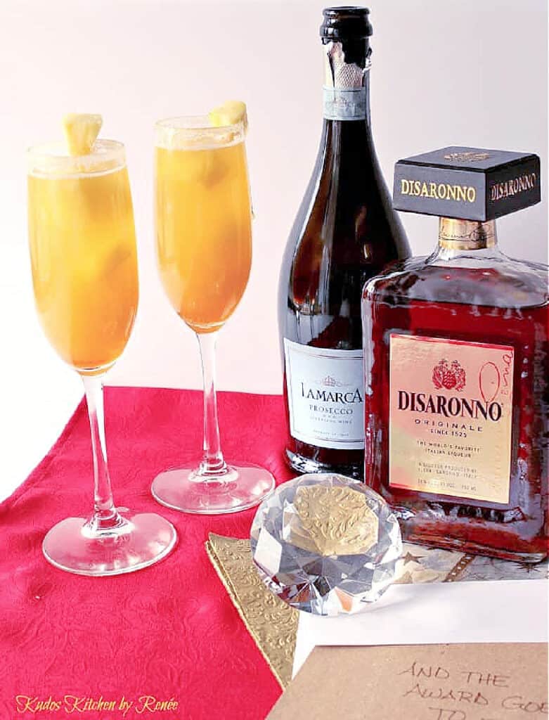 A vertical image of two champagne flutes filled with Golden Statue Prosecco Cocktails on a red and gold napkin and a large diamond for fun.