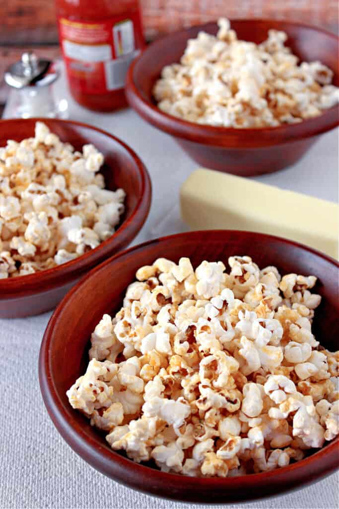 A vertical photo of three bowls filled with Buffalo Popcorn along with a bottle of hot sauce in the background.