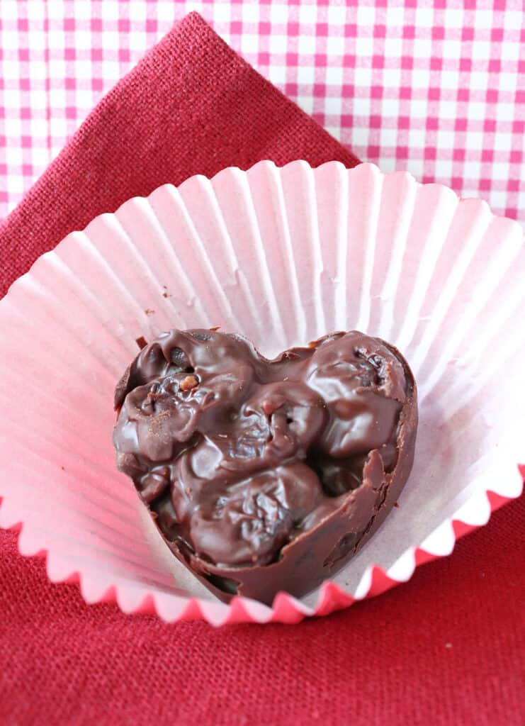 A closeup vertical photo of one Chocolate Covered Dried Cherry Clusters Candy along with a red napkin and a red and white checked background.