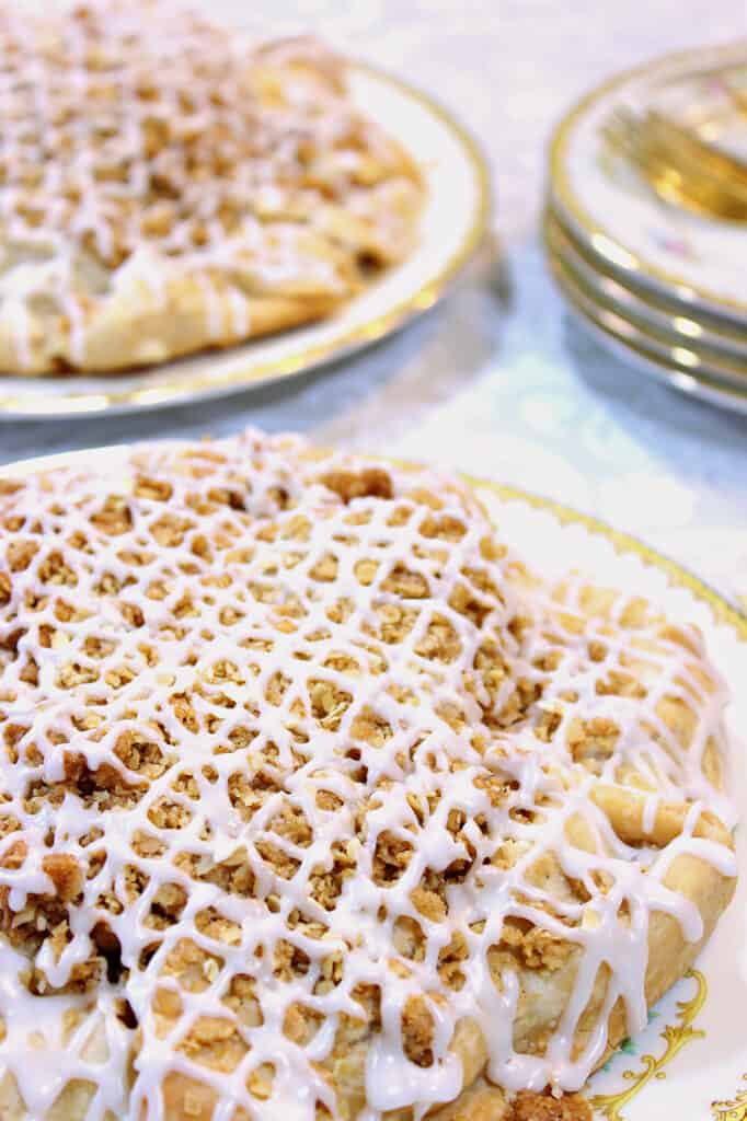 A vertical closeup of an Apple Crumble Crostata in the foreground with a white icing drizzle on top.