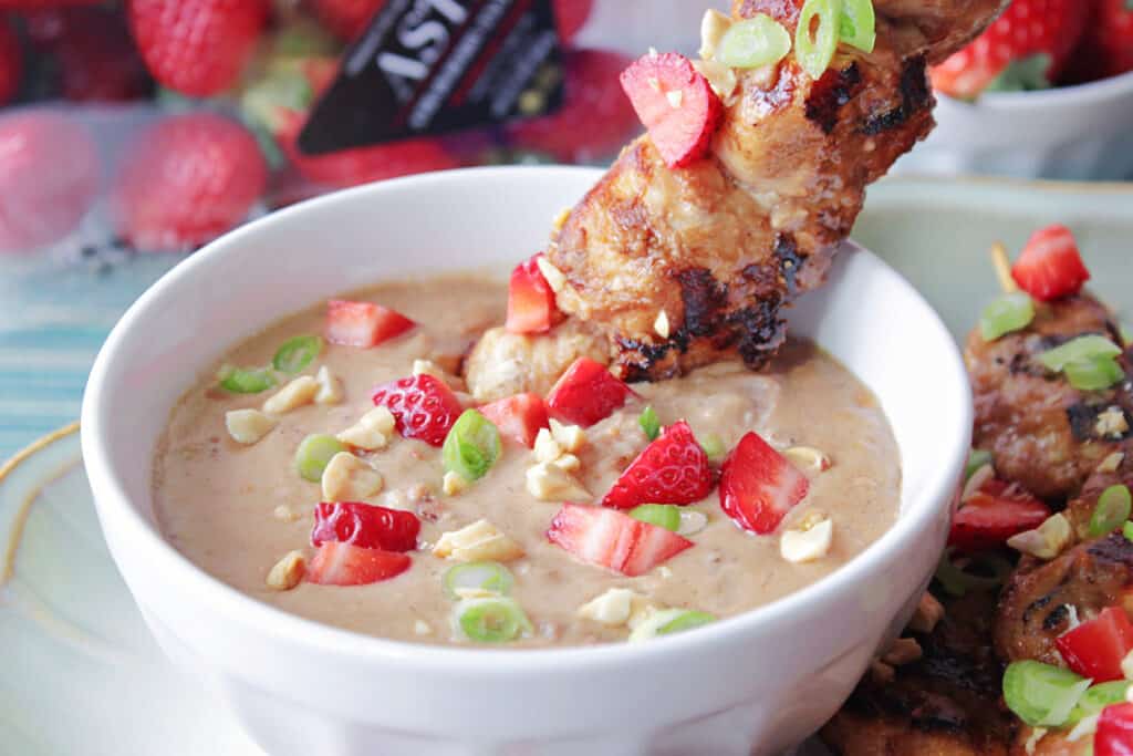 A white bowl filled with Strawberry Satay Sauce along with a chicken tender on a skewer with scallions and peanuts.