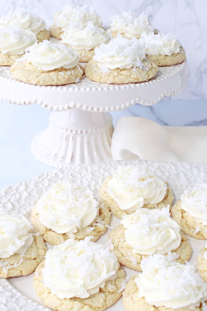 A vertical photo with a white cake stand and white platter filled with Coconut Cake Cookies topped with frosting and coconut.