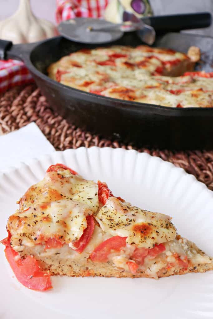 A vertical image of a slice of Deep Dish Garlic Lover's Pizza on a paper plate and a cast iron skillet with the remaining pizza in the background.